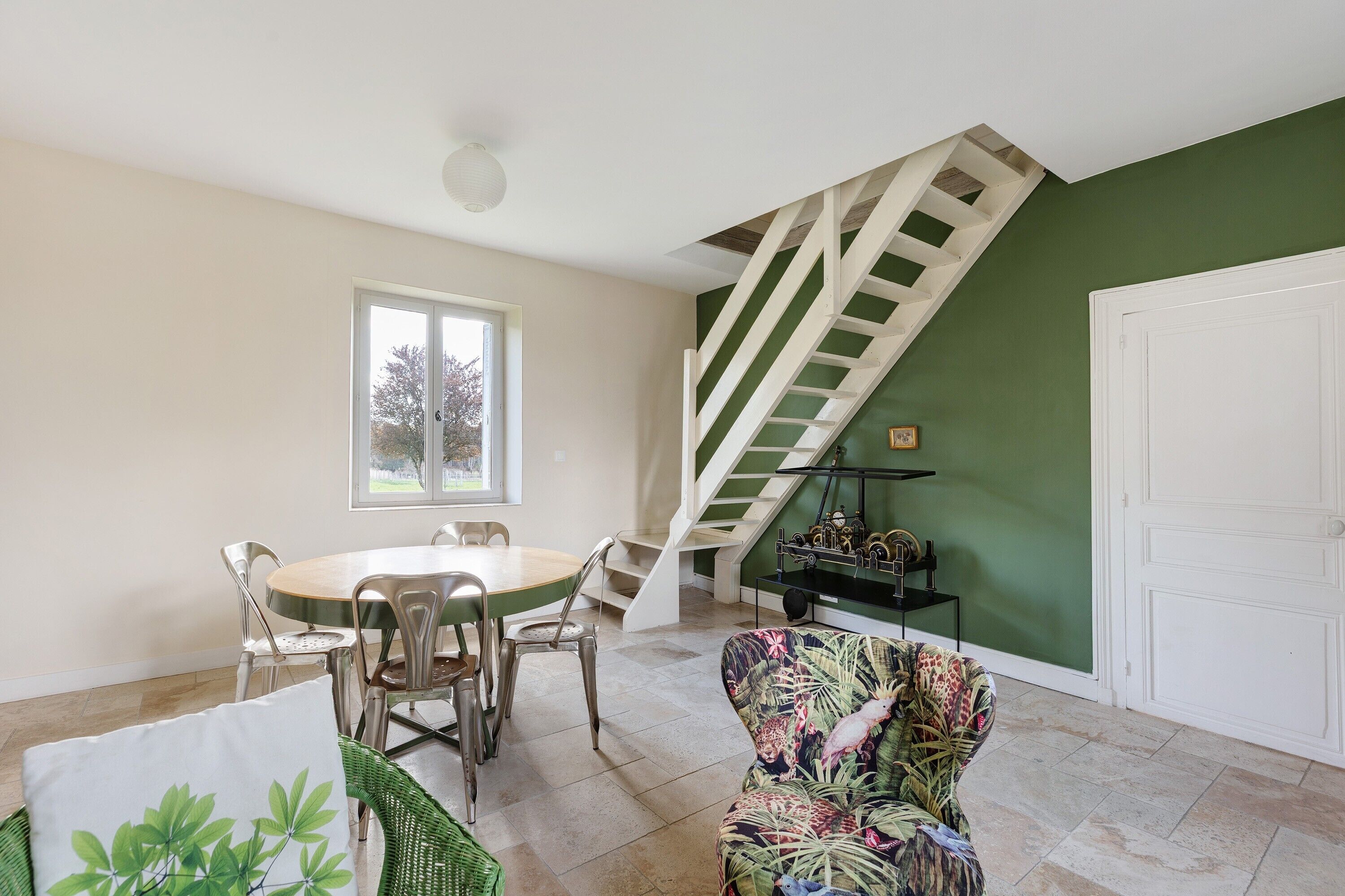 Un coin repas avec une table ronde et des chaises, un escalier avec des rampes vertes et une porte blanche.