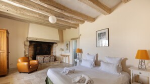 Un dormitorio con una cama grande, chimenea, una silla de madera, una mesita de noche y una lámpara.