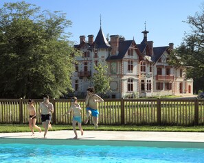 Une piscine entourée d’une clôture en bois, un grand bâtiment doté de nombreuses cheminées et, au loin, une tour.