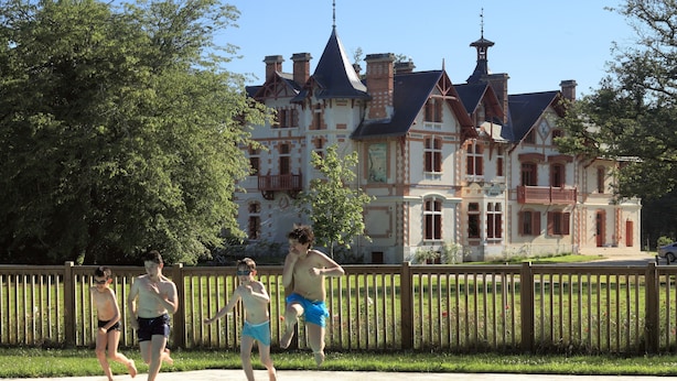 Una piscina con una recinzione in legno sullo sfondo e, in lontananza, un grande edificio storico con numerose canne fumarie e una torre.
