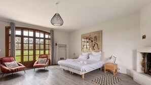 Une chambre avec un grand lit, deux fauteuils, une cheminée et une vue sur l’extérieur.