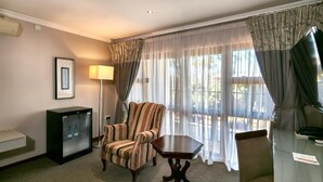 A hotel room with a striped armchair, a small table, a television, a refrigerator, and a window with sheer curtains. - Golden Crystal Hotel (Gaborone)