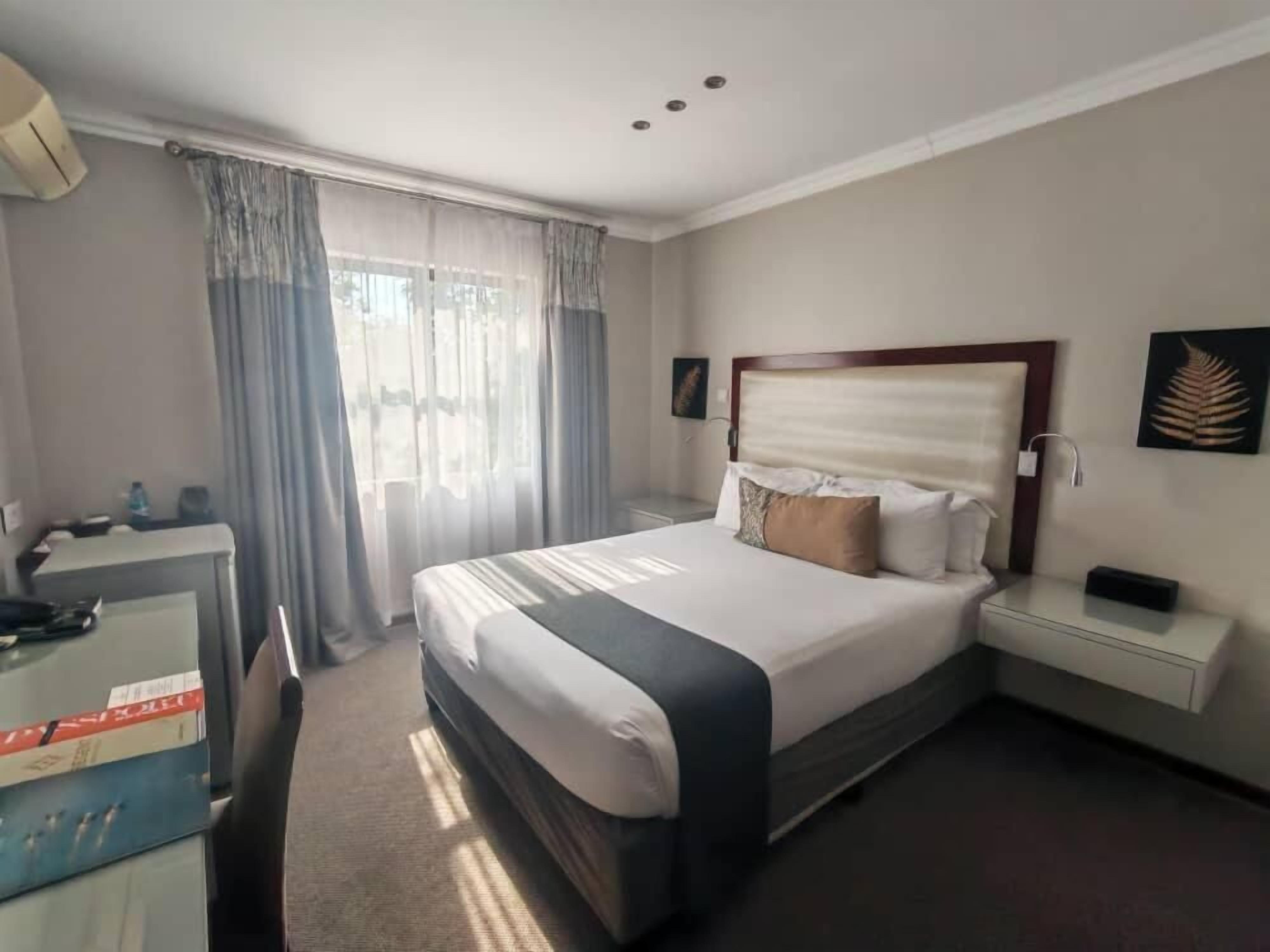 A hotel room with a bed, a desk, a nightstand, and a window with curtains.