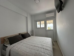 A bedroom with a bed, a window, an air conditioning unit, and a wall-mounted TV. - Loutraki s Hideaway in Loutraki (Loutraki-Agioi Theodoroi)
