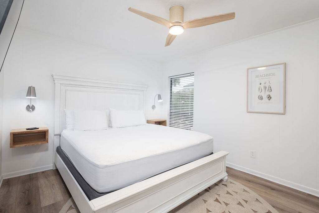 Un dormitorio con cama, ventilador de techo, lámparas de noche, una ventana y un cuadro en la pared.