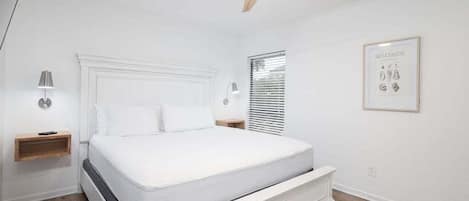 Un dormitorio con cama, ventilador de techo, lámparas de noche, una ventana y un cuadro en la pared.