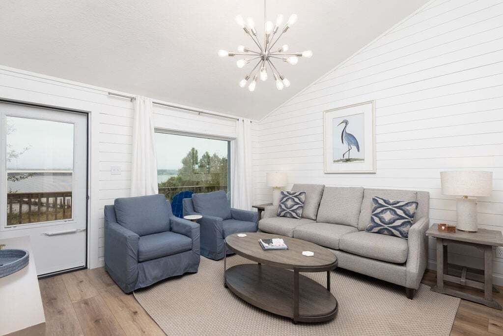 Une pièce avec un canapé gris, des fauteuils bleus, une table basse, un tableau représentant un oiseau encadré et un lustre.