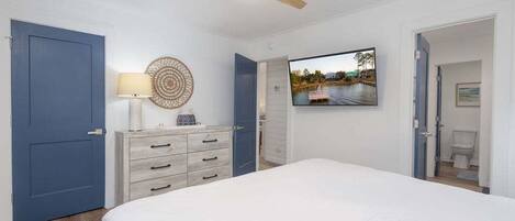 Une chambre à coucher comprenant un lit, un ventilateur de plafond, une télévision, une commode et une porte donnant sur une salle de bain.
