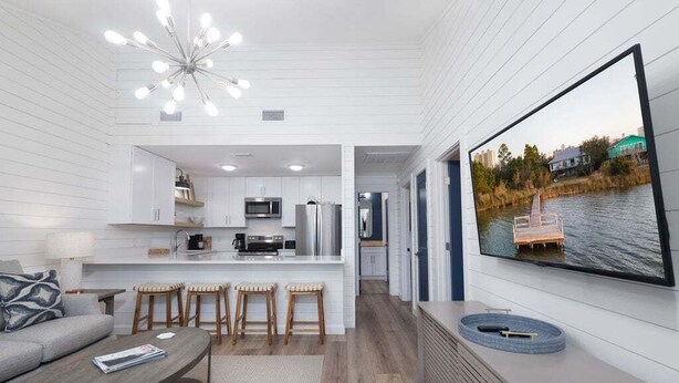 A modern interior with a kitchen, dining area, and a large TV screen displaying a scenic outdoor view. - Sailhouse West 2 Bedroom Duplex (Gulf Shores)