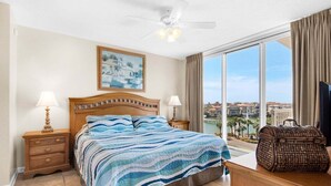 A hotel room with a large bed, two bedside tables with lamps, a ceiling fan, a large window with a view, and a wicker trunk. - Terrace At Pelican Beach 401 3 Bedroom Home (Destin)