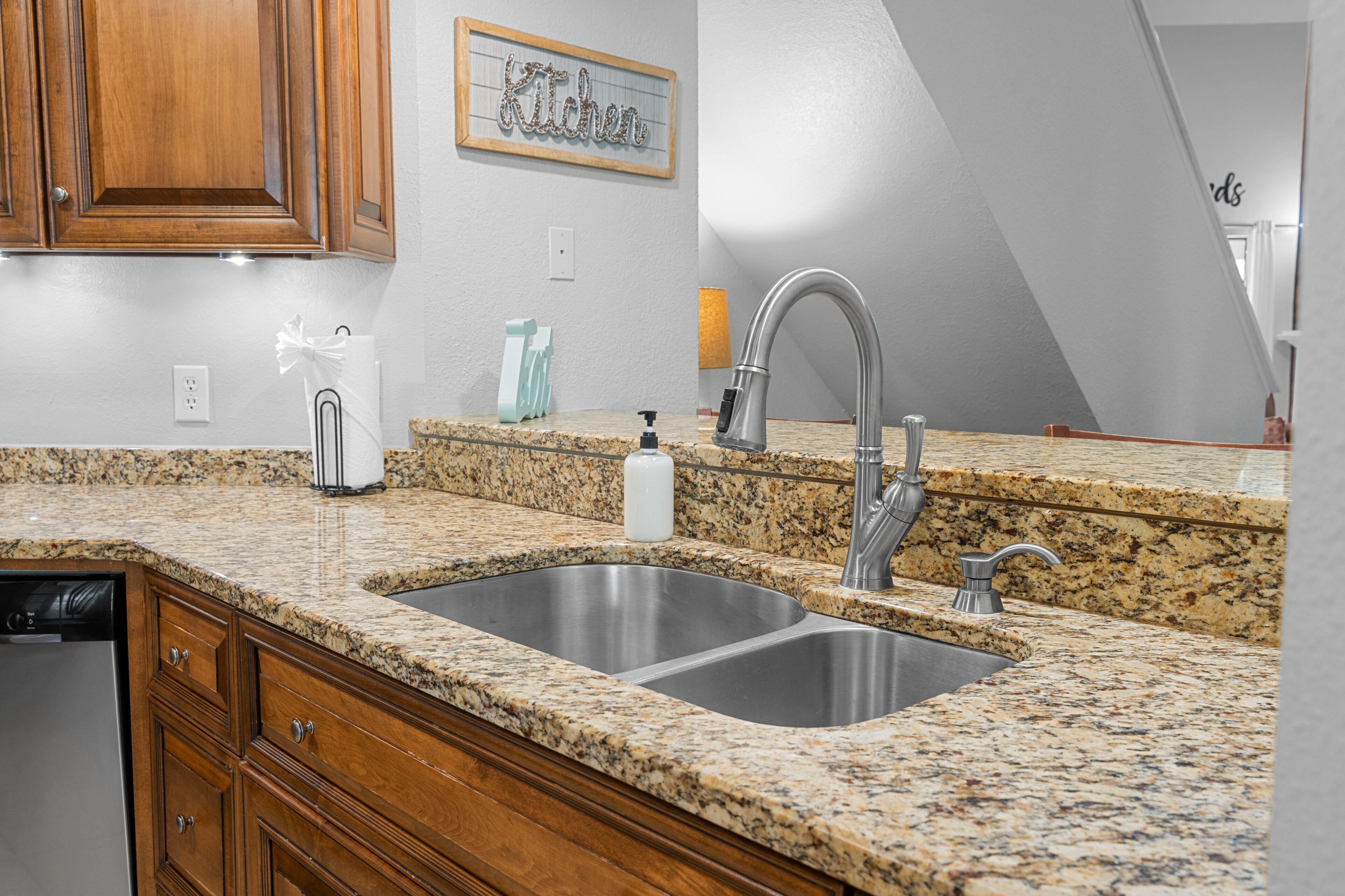 A kitchen with granite countertops, stainless steel sink, wooden cabinets, and a framed 'Kitchen' sign on the wall.