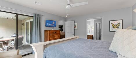 Un dormitorio con cama, ventilador de techo, televisor y una puerta que da a otra habitación.