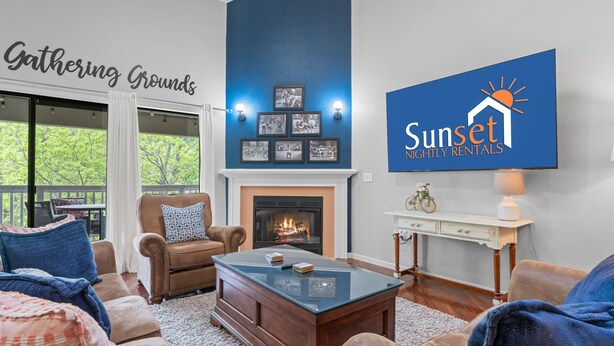 A living room with a fireplace, a sofa, a coffee table, and a television displaying a logo. - Pr66-8 4 Bedroom Condo (Branson)