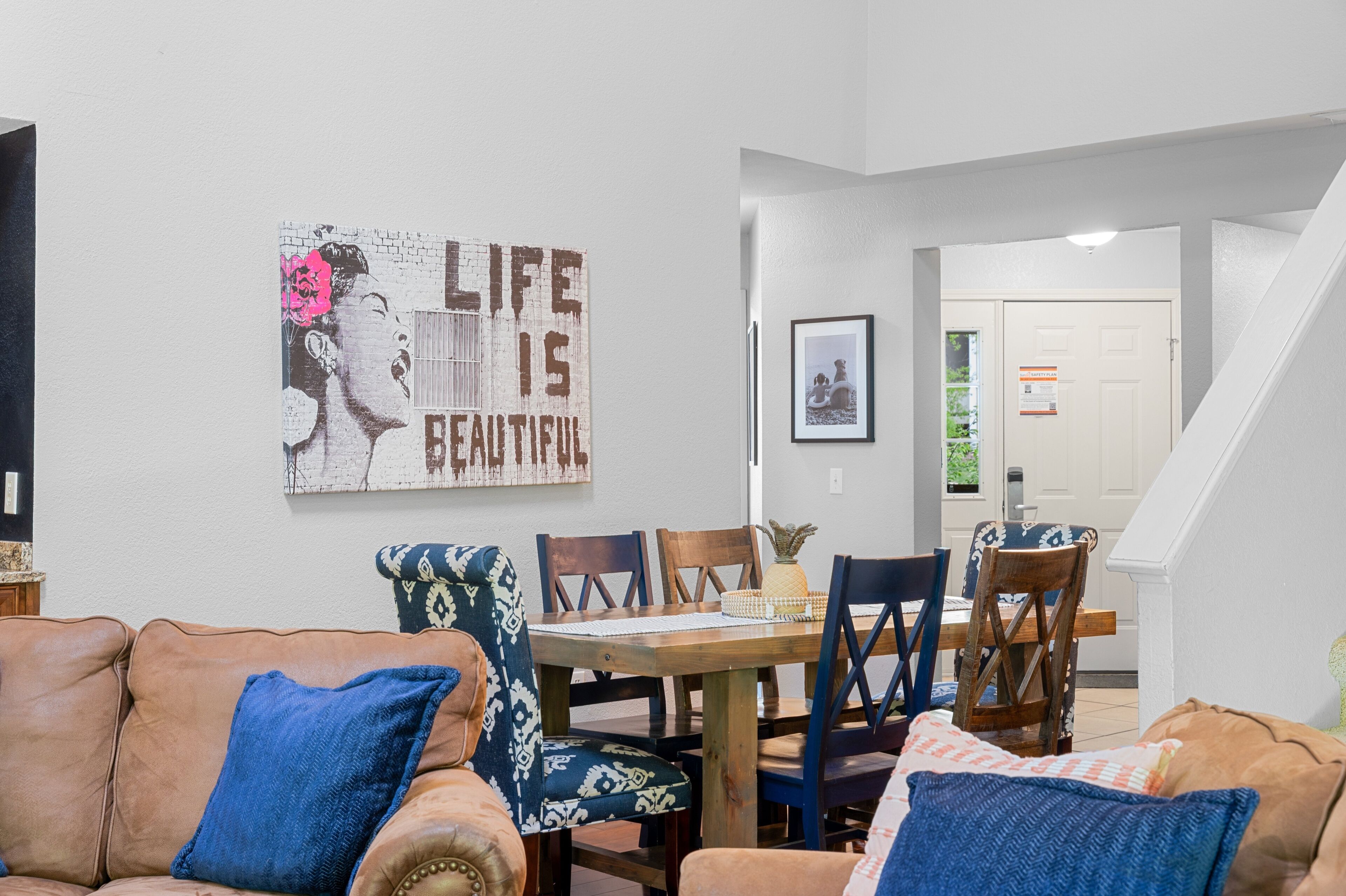Un comedor con una mesa y sillas de madera, una pared con un cartel, y una escalera que conduce a otra habitación.