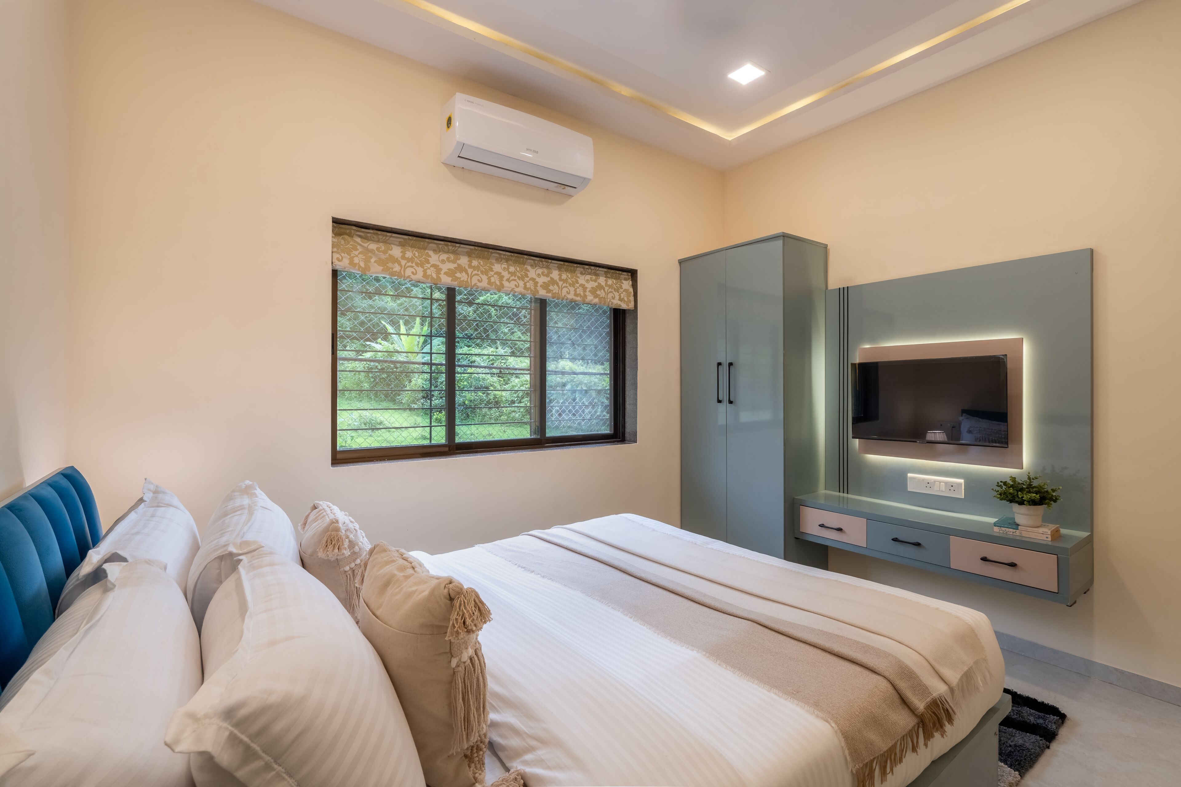 A bedroom with a bed, a TV, a wardrobe, and a window with a view of greenery.
