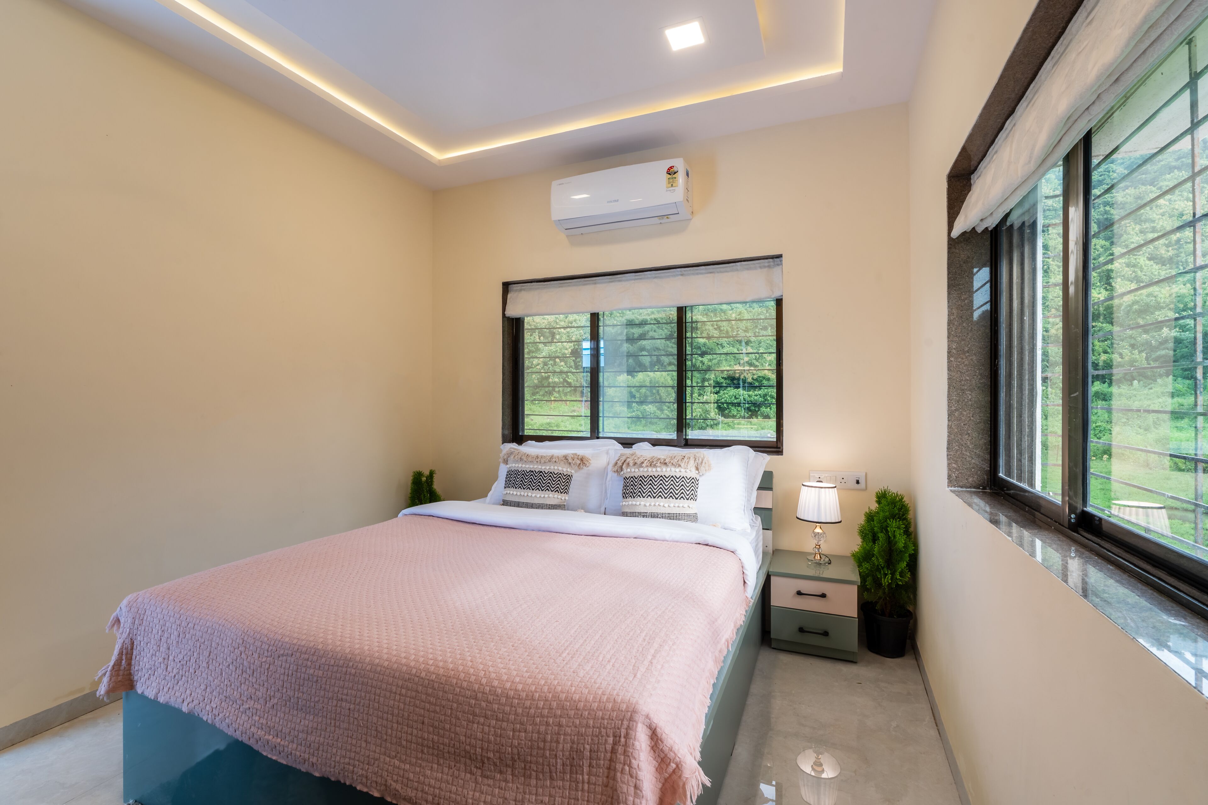 A modern bedroom with a bed, bedside table, lamp, air conditioning unit, and a window with a view of greenery.