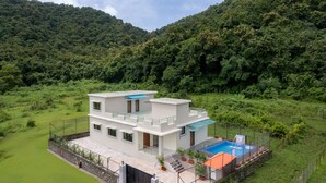 Ein modernes, zweistöckiges Haus mit Swimmingpool, umgeben von üppiger Vegetation und Hügeln.