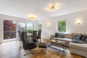 A modern living room with a sofa, chairs, a coffee table, and large windows offering a view of trees and a balcony. - Chalet on Lake Diemelsee with Lake View in the Sauerland (Diemelsee)