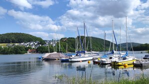 Un lago tranquilo con varias embarcaciones amarradas, rodeado de vegetación y un pequeño pueblo al fondo.