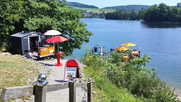 Una zona junto al lago con un muelle pequeño, un buzón rojo con la marca Coca-Cola y una zona de barbacoa.