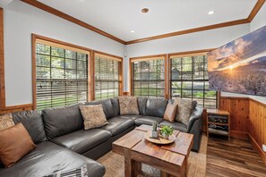 Una sala de estar moderna con un sofá modular, piso de madera, ventanas amplias con persianas y un cuadro en la pared.