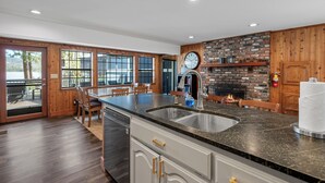 A kitchen with a large island, a brick fireplace, and a view through the glass doors. - Lakefront Escape with Private Dock, Hot Tub, AC, EV charger, & Game Room (Big Bear Lake)