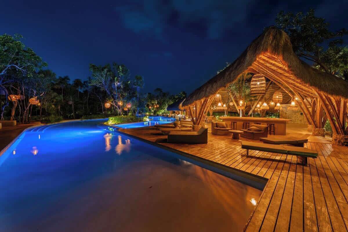 Un espace aménagé au bord de la piscine, avec une terrasse en bois, une structure à toit de chaume et des sièges d’extérieur.