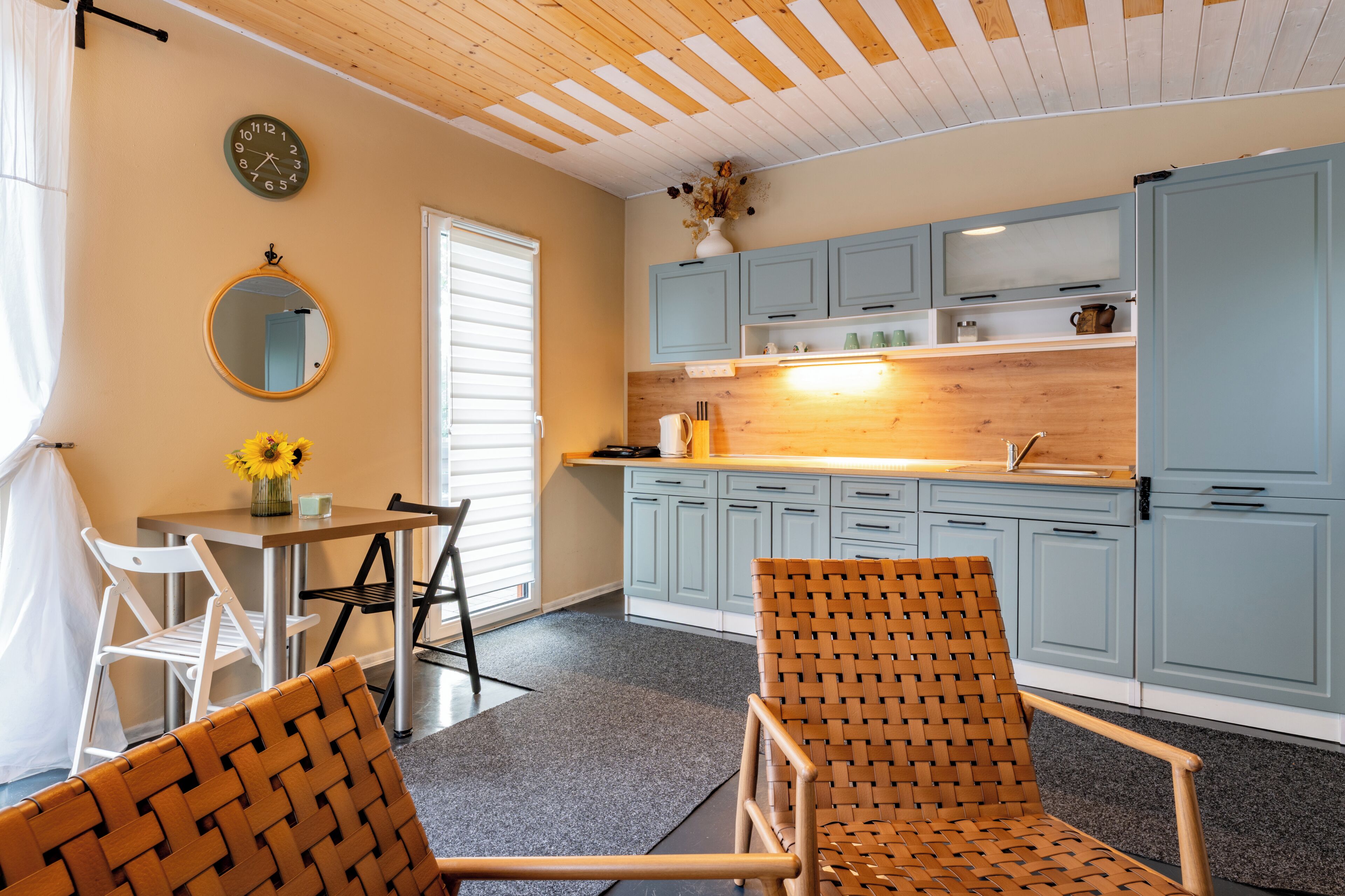 A kitchen with light blue cabinets, a wooden countertop, a round mirror, and a dining area with two wicker chairs and a table.