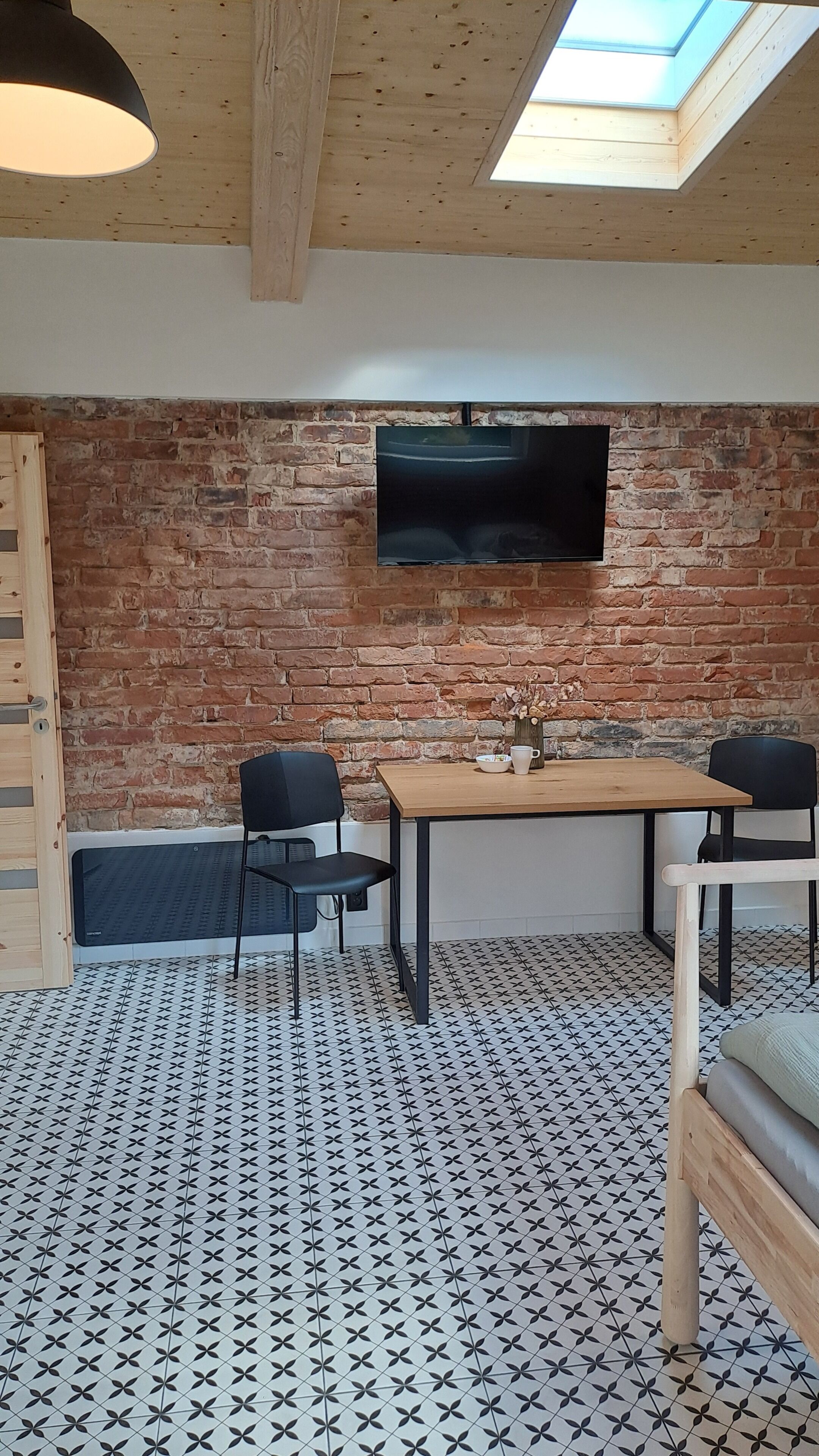 Een kamer met een bakstenen muur, een flatscreen televisie boven een houten tafel, twee stoelen en een kamer met een patroon vloer.