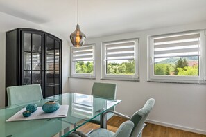 A modern room with a glass table, chairs, a black cabinet, and large windows with a view of greenery.