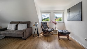 A loft-style room with a sloped ceiling, a gray sofa, a leather chair, a tripod-mounted lamp, and a painting of a bridge. - Rural, Quiet Apartment (Rosengarten)