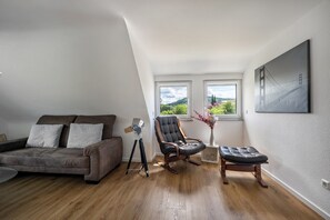 A loft-style room with a sloped ceiling, a gray sofa, a leather chair, a tripod-mounted lamp, and a painting of a bridge.