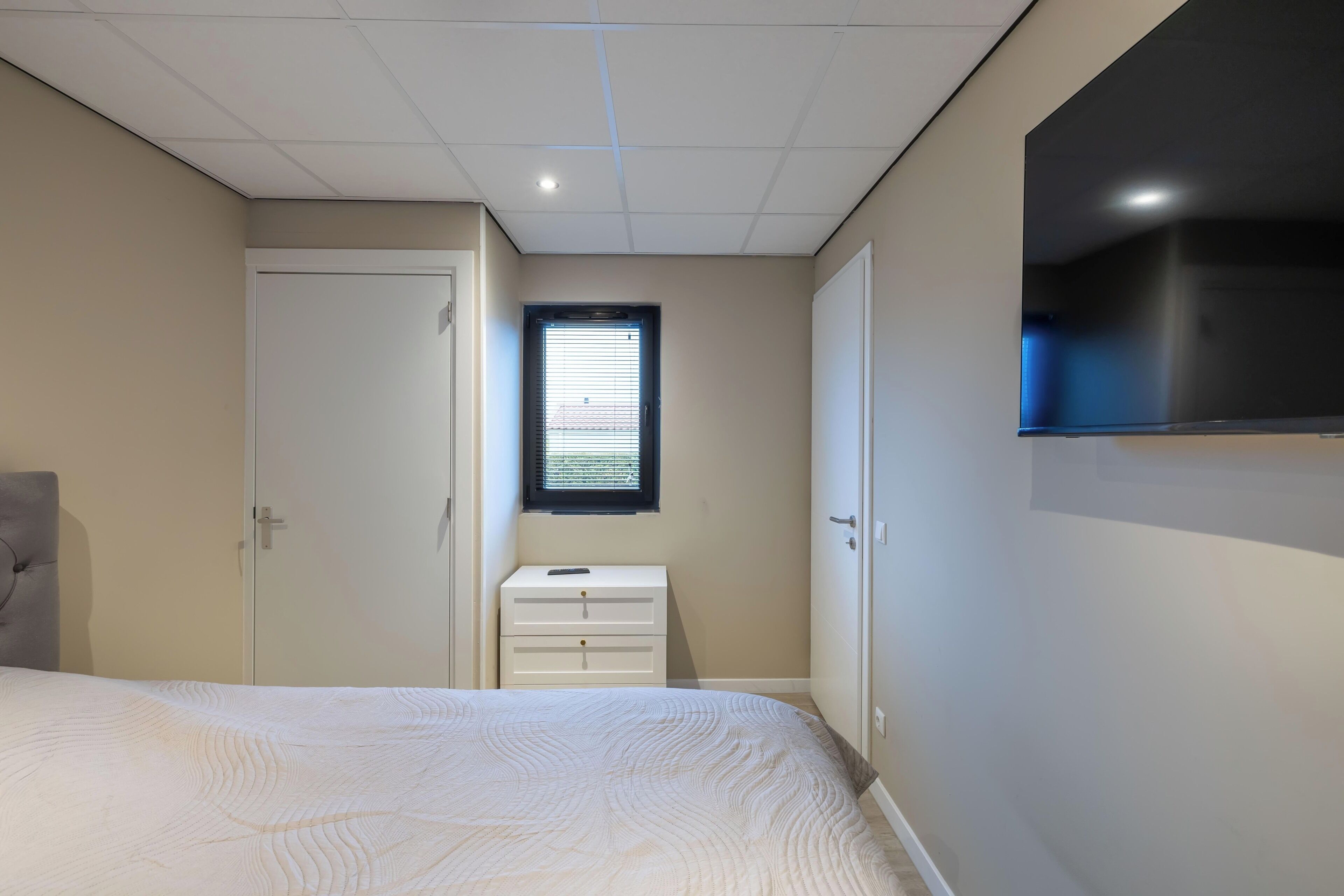 A bedroom with a bed, a wardrobe, a dresser, a window, and a TV mounted on the wall.