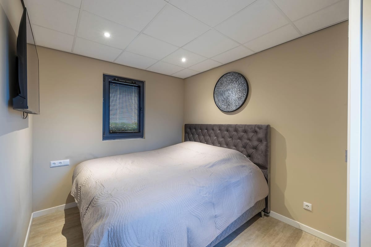 A bedroom with a bed, a wall-mounted TV, a window with blinds, and a circular wall decoration.