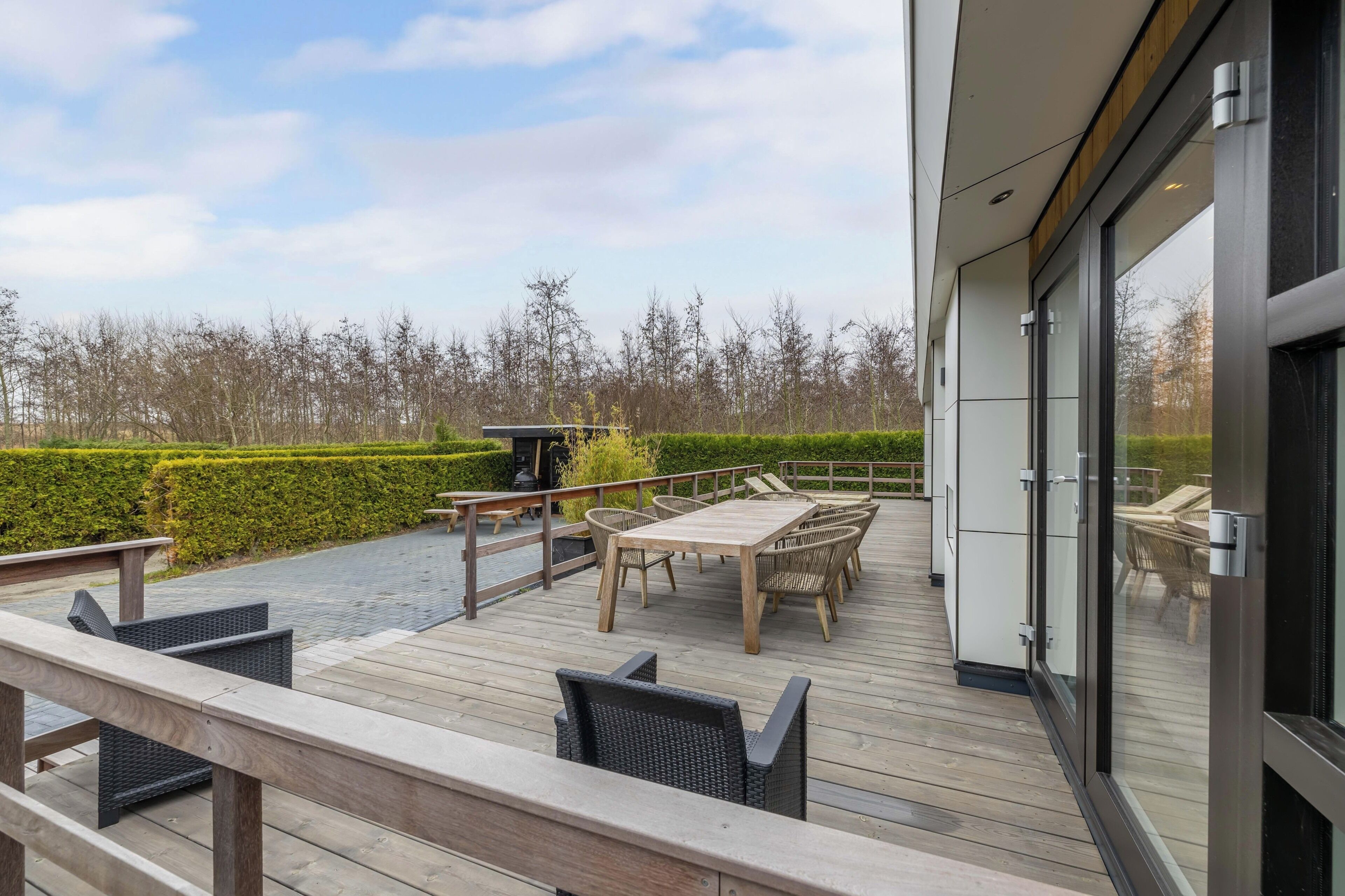 Une terrasse dotée de mobilier d’extérieur, d’une table et de chaises, donnant sur un espace paysager agrémenté d’une haie et d’arbres.