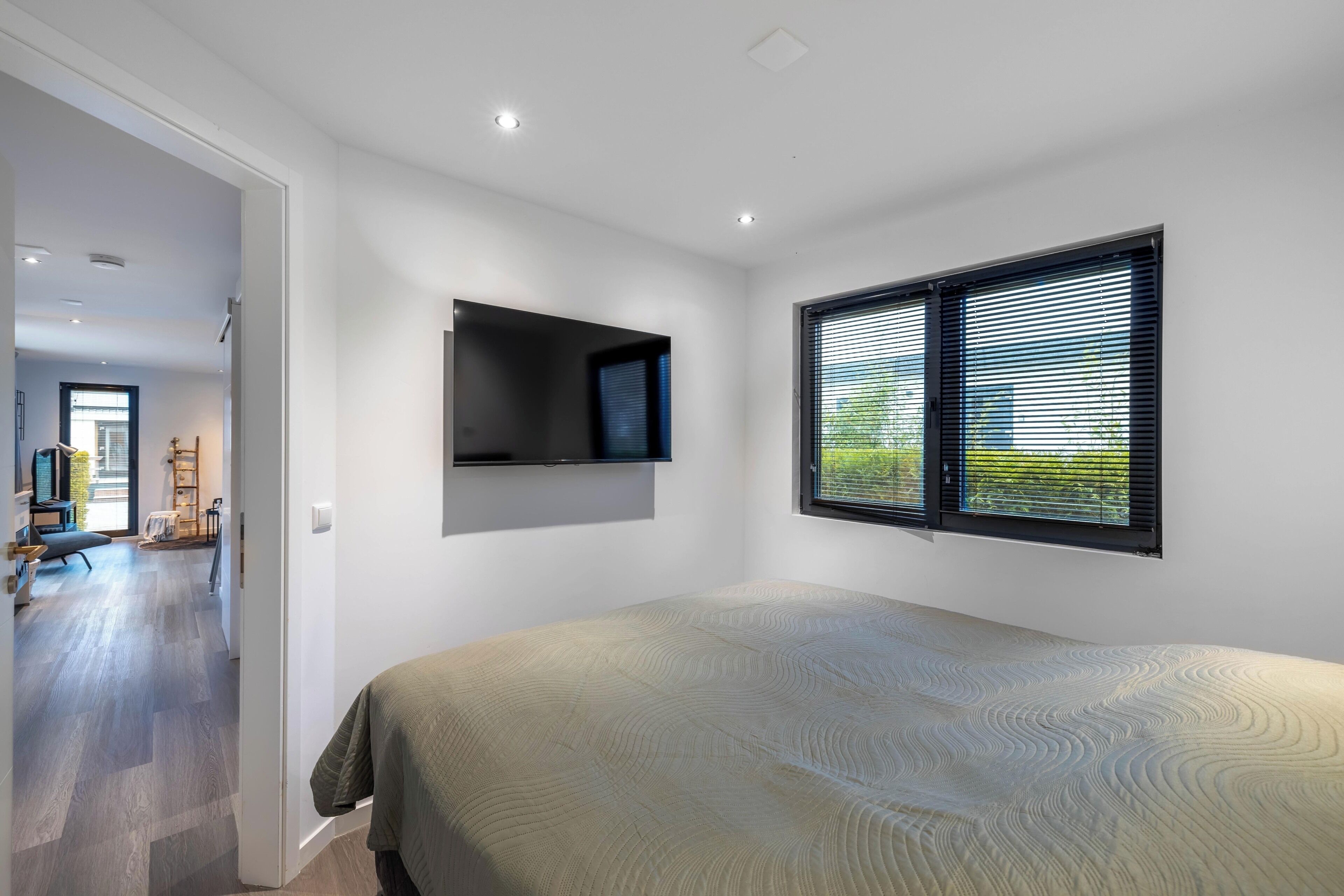 A bedroom with a bed, a mounted TV, and a window with blinds.