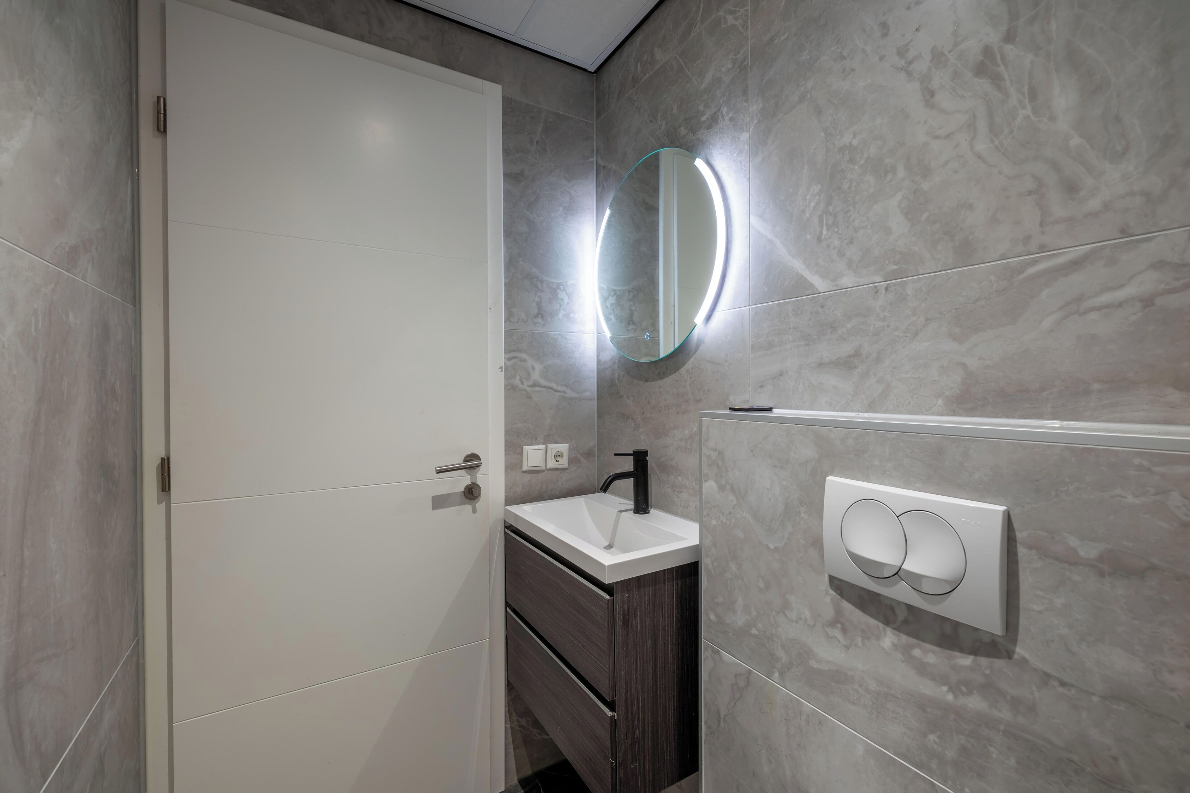 A bathroom with a white sink, dark wooden cabinet, and a wall-mounted mirror with a light.