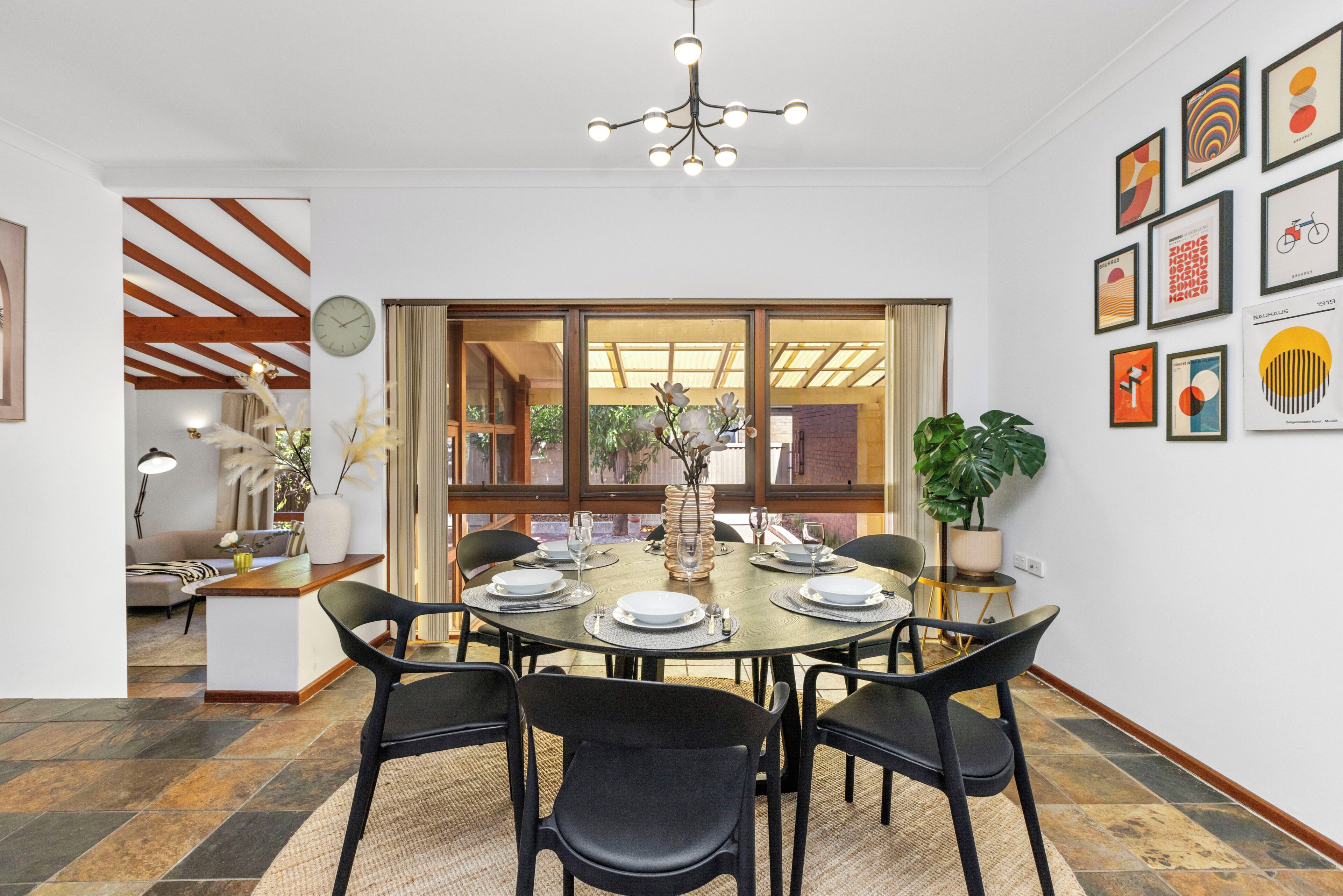 A dining area with a round table set for six, surrounded by black chairs, with a chandelier above and a large window with curtains.