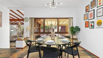 A dining area with a round table set for six, surrounded by black chairs, with a chandelier above and a large window with curtains.