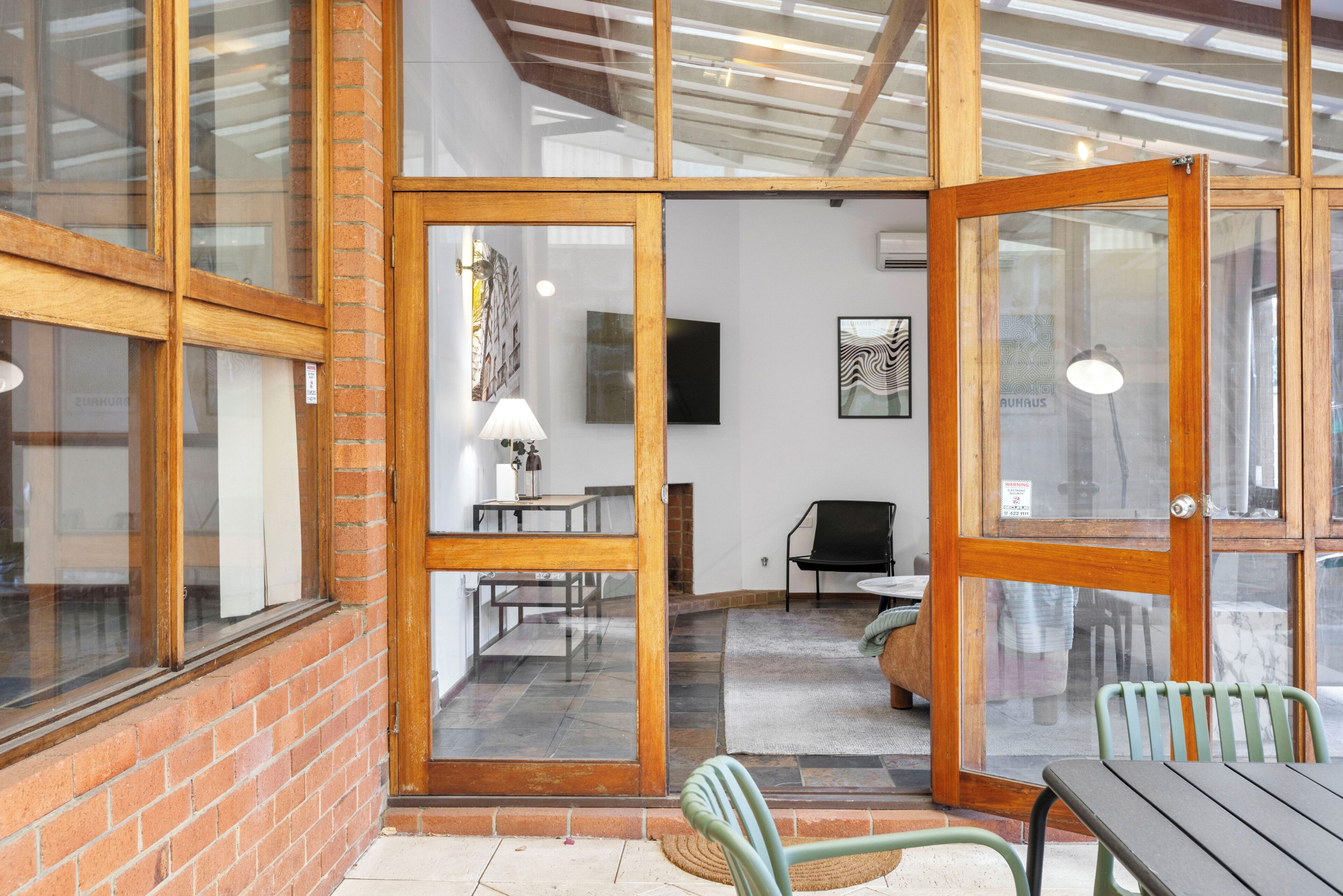 A room with a brick wall, wooden-framed glass doors, a flat-screen TV, a lamp, and a chair.
