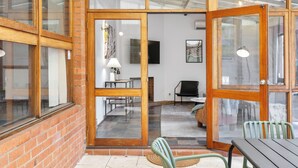 Una habitación con pared de ladrillo, puertas de vidrio con marco de madera, un televisor de pantalla plana, una lámpara y una silla.
