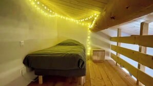 Une petite chambre mansardée, chaleureuse et accueillante, avec un plancher en bois, un lit simple et des guirlandes lumineuses au plafond.