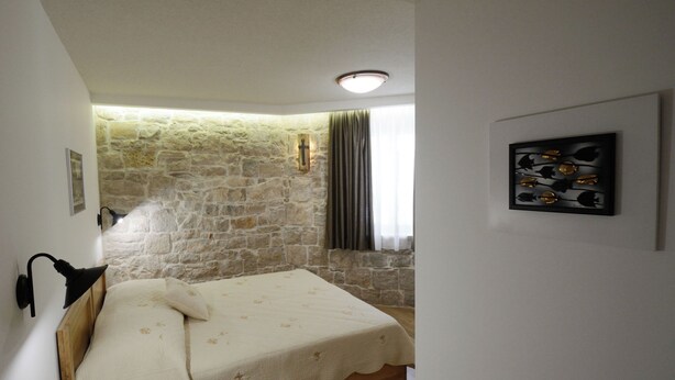 Une chambre avec un mur en pierre, un lit recouvert d’une courtepointe blanche, une tête de lit en bois, une lampe fixée au mur et un tableau encadré accroché au mur.