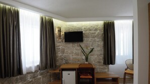Une pièce avec un mur en pierre, un téléviseur à écran plat fixé au-dessus d’un bureau en bois, un petit réfrigérateur, une chaise et une fenêtre avec des rideaux.