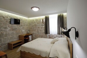 A bedroom with a stone wall, a flat-screen TV, a bed with a white quilt, a wooden bench, and a desk. - Apartment and Rooms Desideria (Split)