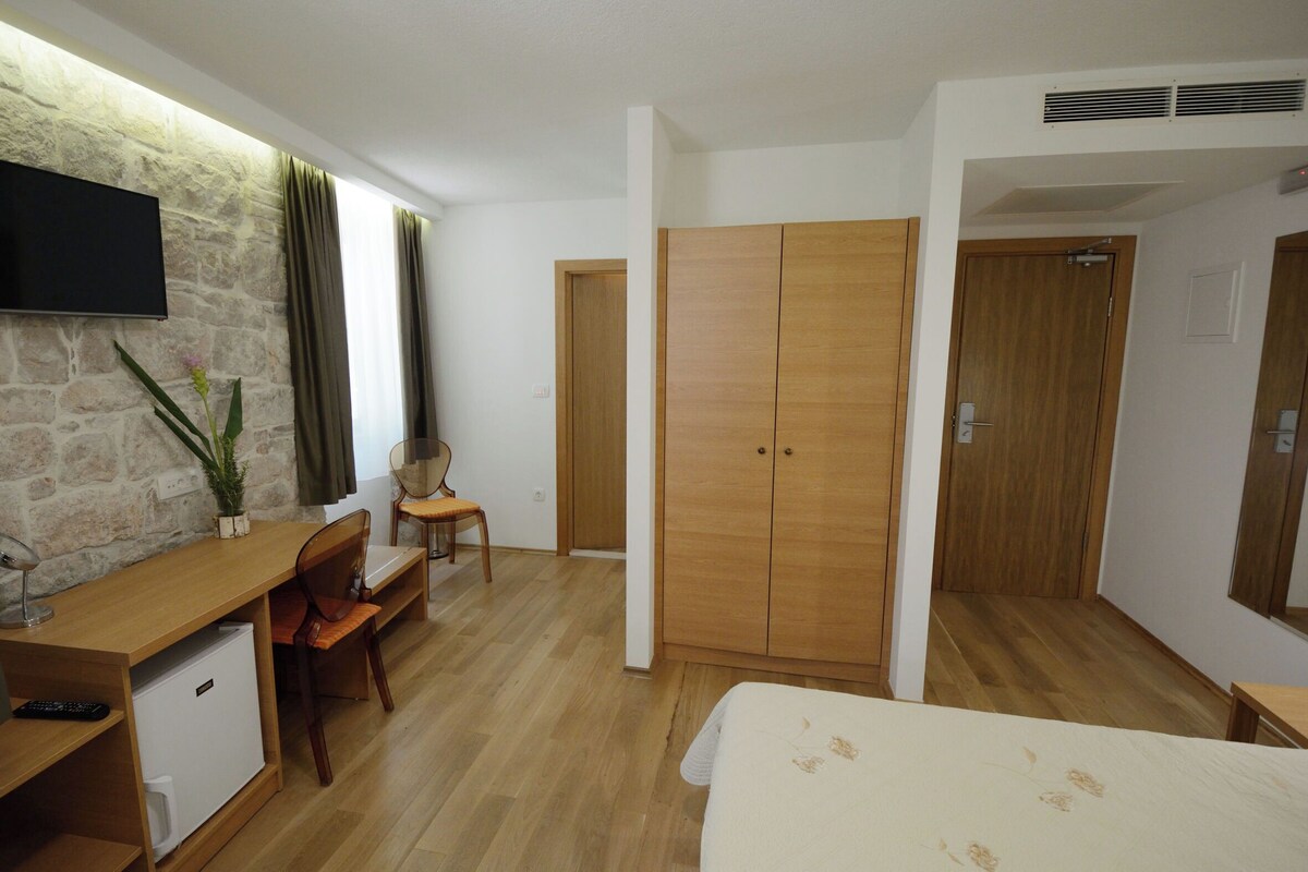 Ein Hotelzimmer mit einem Bett, einem Schreibtisch mit Stuhl, einem kleinen Kühlschrank, einem an einer Steinwand befestigten Fernseher und Holzschränken.