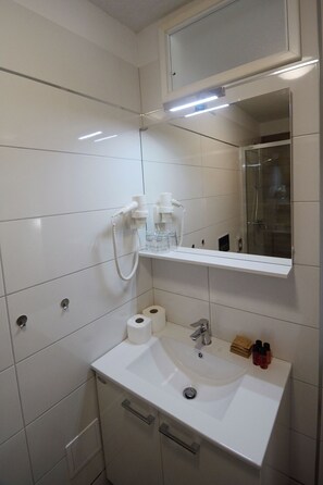 A bathroom with a white sink, toilet, and shower. There is a mirror above the sink and a shelf with toiletries. - Apartment and Rooms Desideria (Split)