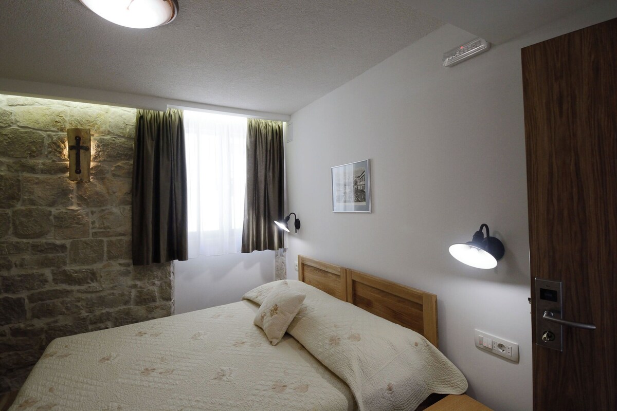 Ein Schlafzimmer mit Steinwand, einem Bett mit Kopfteil, einem Fenster mit Vorhängen, einer Wandleuchte, einer Tür und einem kleinen Bild an der Wand.