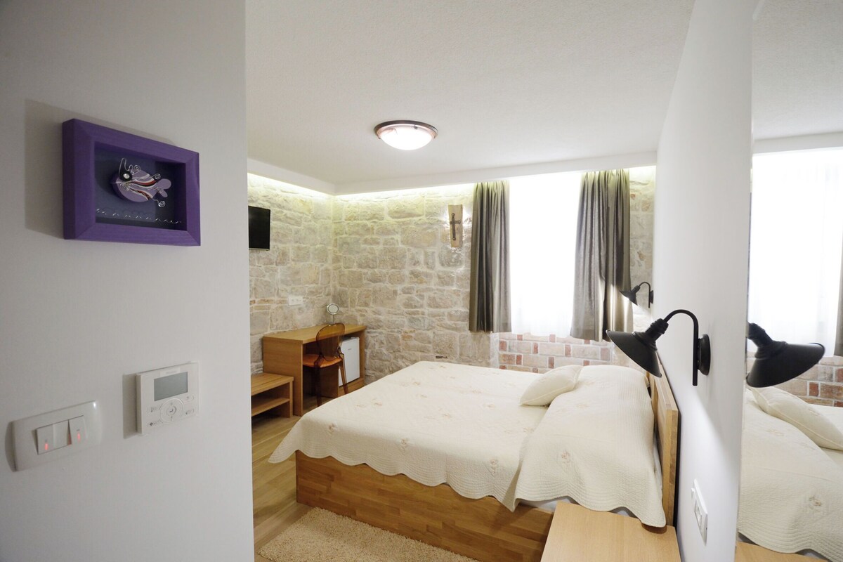 Ein Schlafzimmer mit einer Steinwand, einem Bett mit weißer Bettdecke, einem Schreibtisch mit Stuhl und einem Bild in einem violetten Rahmen an der Wand.