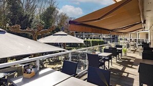 Ein Sitzbereich im Freien mit Tischen und Stühlen, beschattet von großen Sonnenschirmen, umgeben von viel Grün.